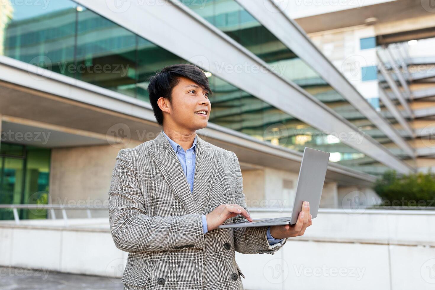 A young Asian man stands outside a contemporary building, engaged in work on his laptop. The setting highlights the blend of nature and modern design around him. photo