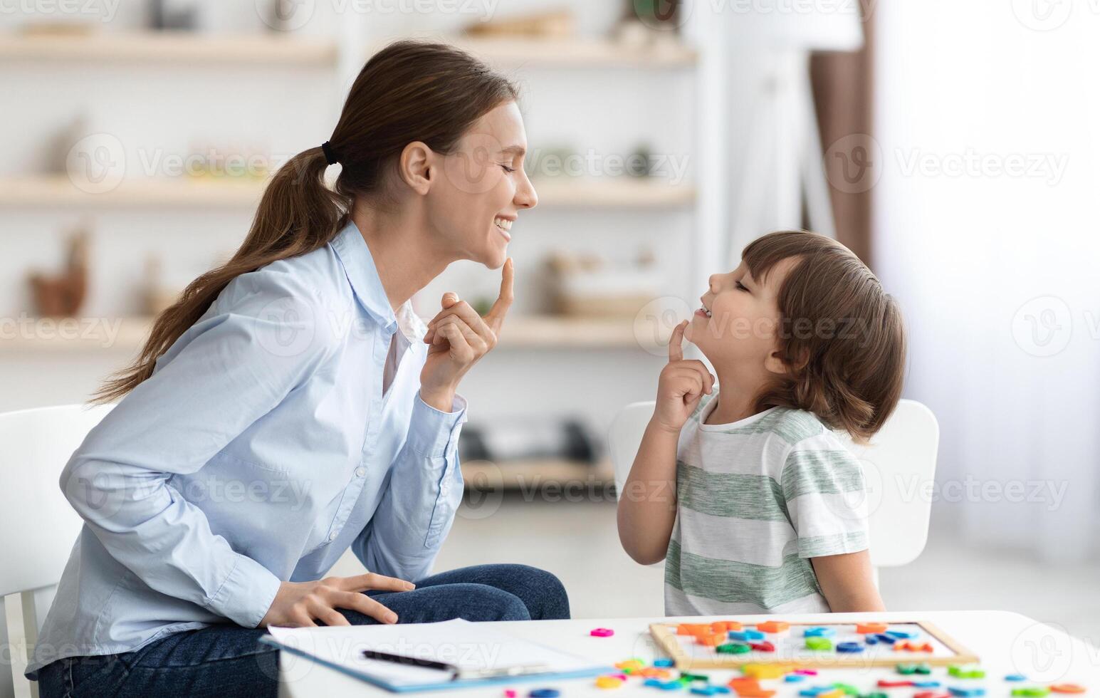 Speech training for kids. Professional woman specialist training with little boy at cabinet, teaching him right articulation exercises, side view photo