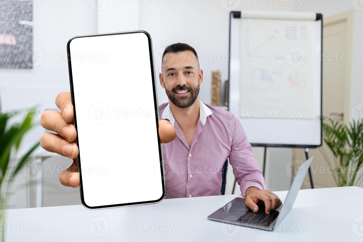 A smiling man sits at a desk in a contemporary office, holding a blank smartphone screen toward the viewer while working on a laptop, highlighting a friendly and tech-savvy atmosphere. photo