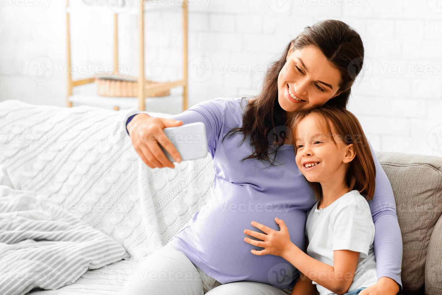 Happy expecting woman taking selfie with her little daughter while sitting on sofa at home, free space photo
