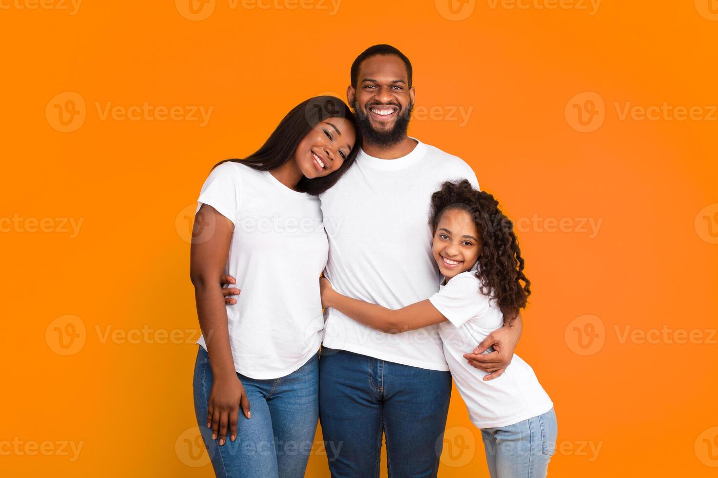 Healthy Relationship. Portrait of cheerful African American family of three people of color hugging, standing isolated on yellow orange studio wall, posing looking at camera, wearing white t-shirts photo