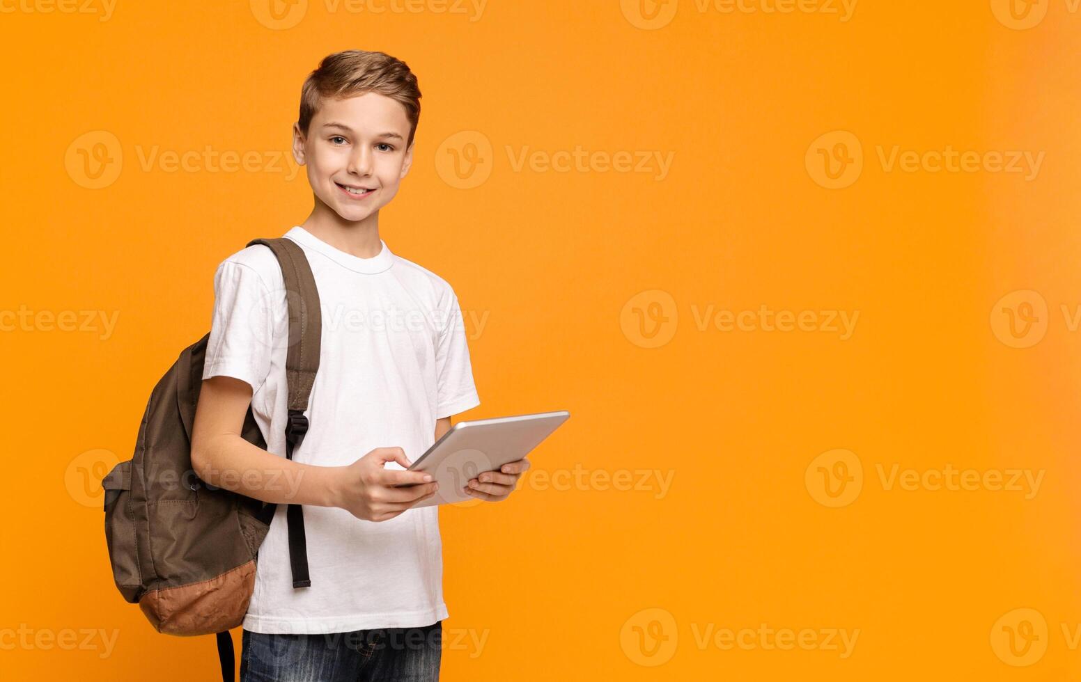 Education and modern technologies. Schoolboy with backpack using tablet computer and smiling on orange background, empty space photo