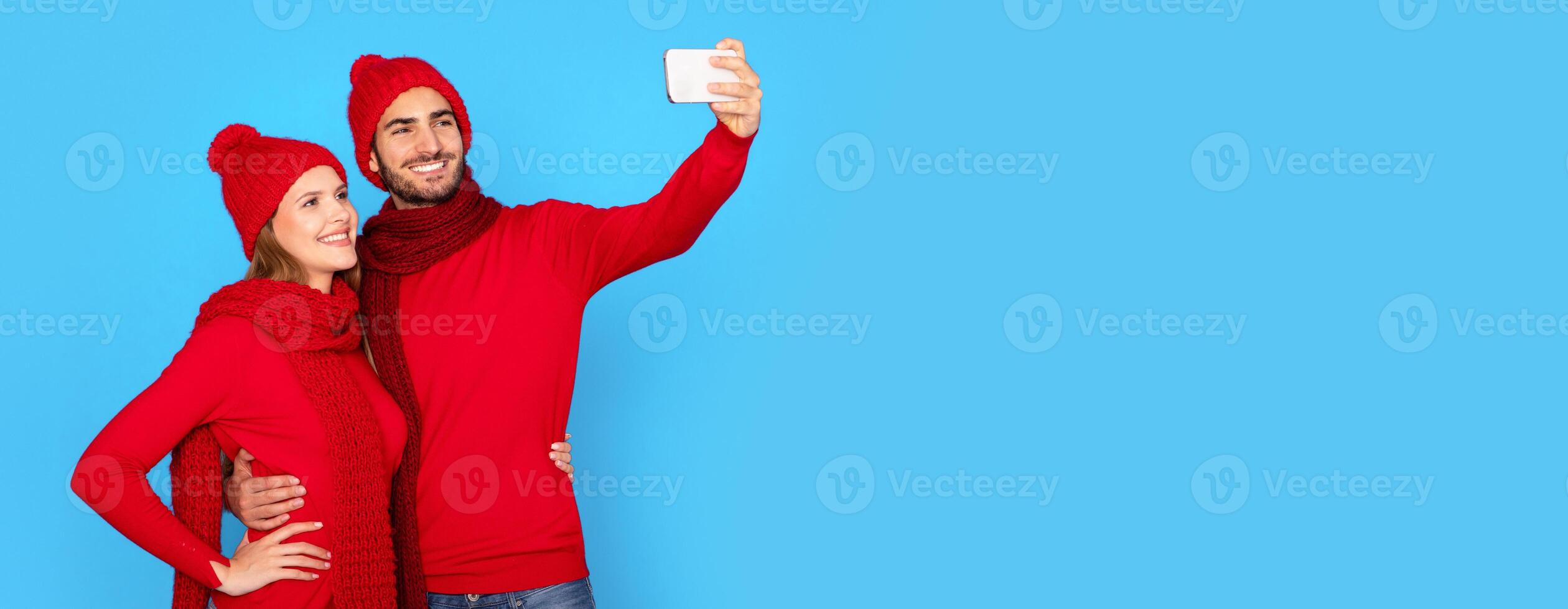 Happy Young Couple Wearing Knitted Hats Taking Selfie On Smartphone, Cheerful Millennial Spouses Making Photos On Mobile Phone And Having Fun Together, Posing Over Blue Studio Background, Copy Space