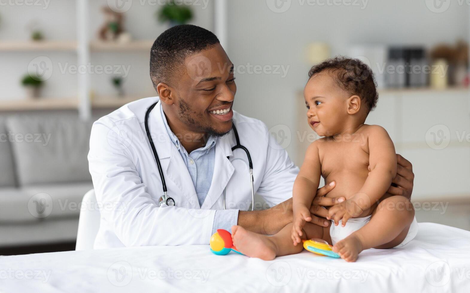 Child's Healthcare Concept. Portrait Of Smiling Black Doctor Making Check Up For Little Infant Baby Boy, Cute Toddler Child In Diaper Having Appointment At Pediatrician's Office, Free Space photo