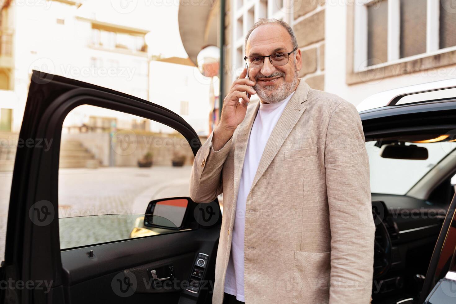 A well-dressed man stands beside his parked car in an urban setting, smiling while engaged in a phone conversation. The environment reflects a charming city atmosphere. photo