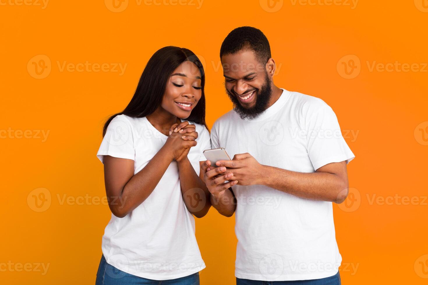 Excited black couple looking at cell phone screen, using new cool application, standing isolated on orange studio wall. Joyful African American man and woman using smartphone together, sharing media photo