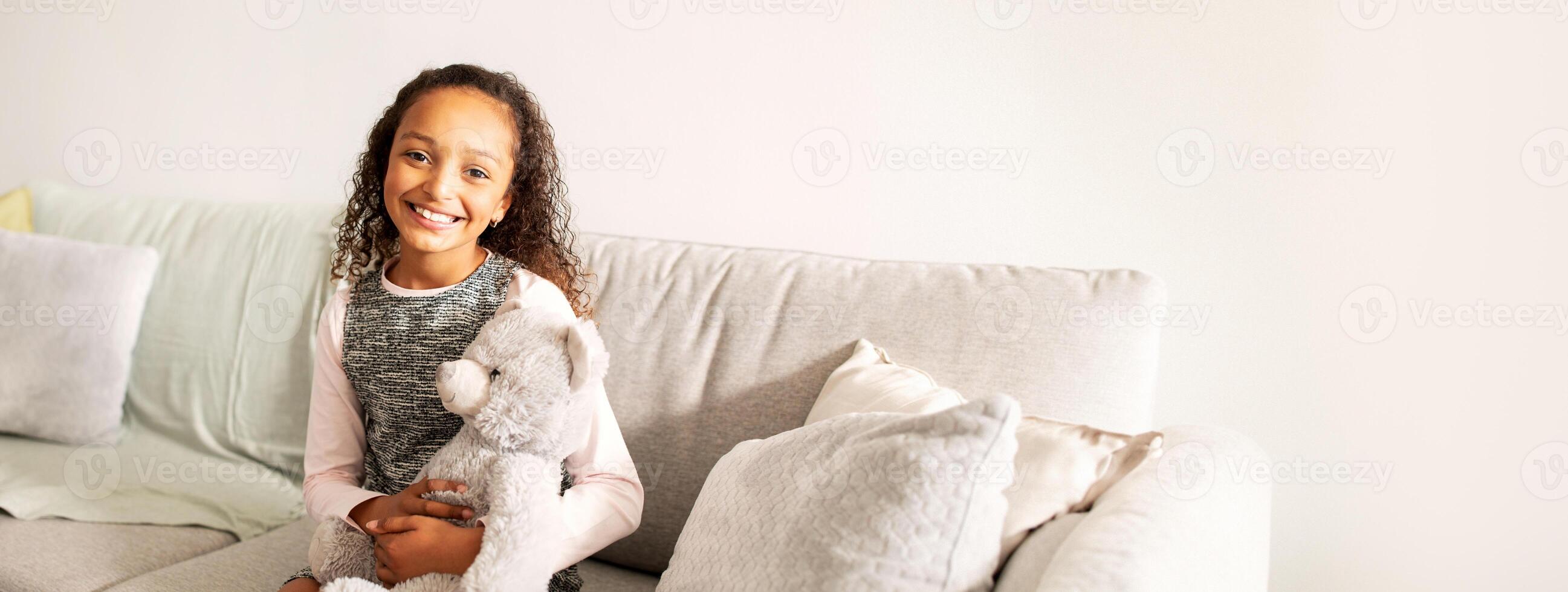 A cheerful black girl sits on a light-colored couch, holding her teddy bear and smiling warmly. The setting is bright and inviting, enhancing her joyful demeanor. photo