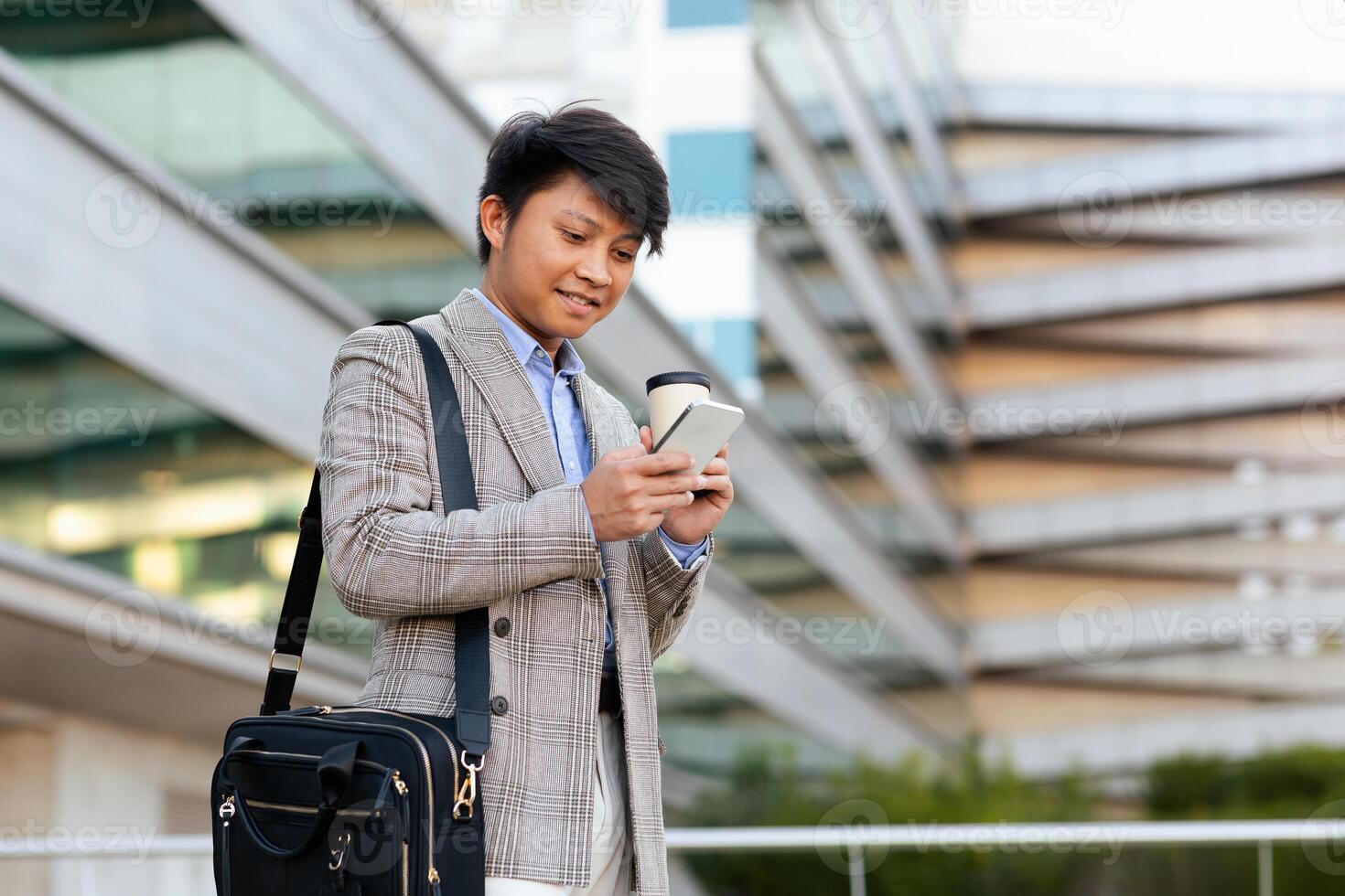 A young Asian businessman in a plaid suit and carrying, a bag smiles as he checks his smartphone outside a contemporary office building during the day. photo