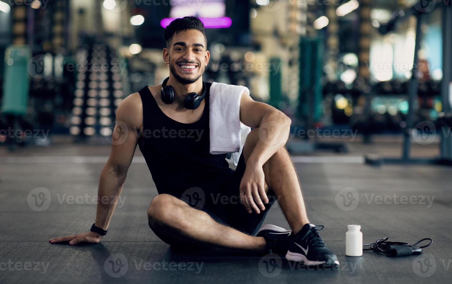Young Arab Male Athlete Relaxing On Floor After Training At Gym, Smiling Middle Eastern Man Resting Next To Blank Container With Supplement Pills, Enjoying Bodybuilding And Healthy Lifestyle photo