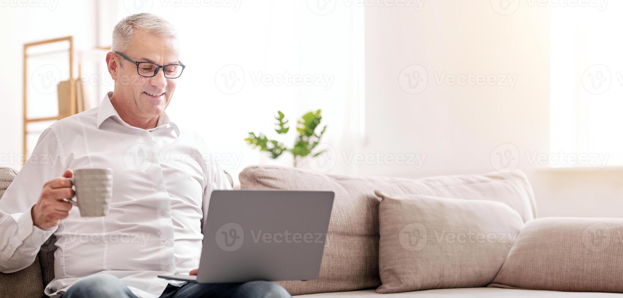 In The Morning. Mature Man Holding Computer On His Lap, Having Tea Sitting On Sofa At Home, Free Space photo