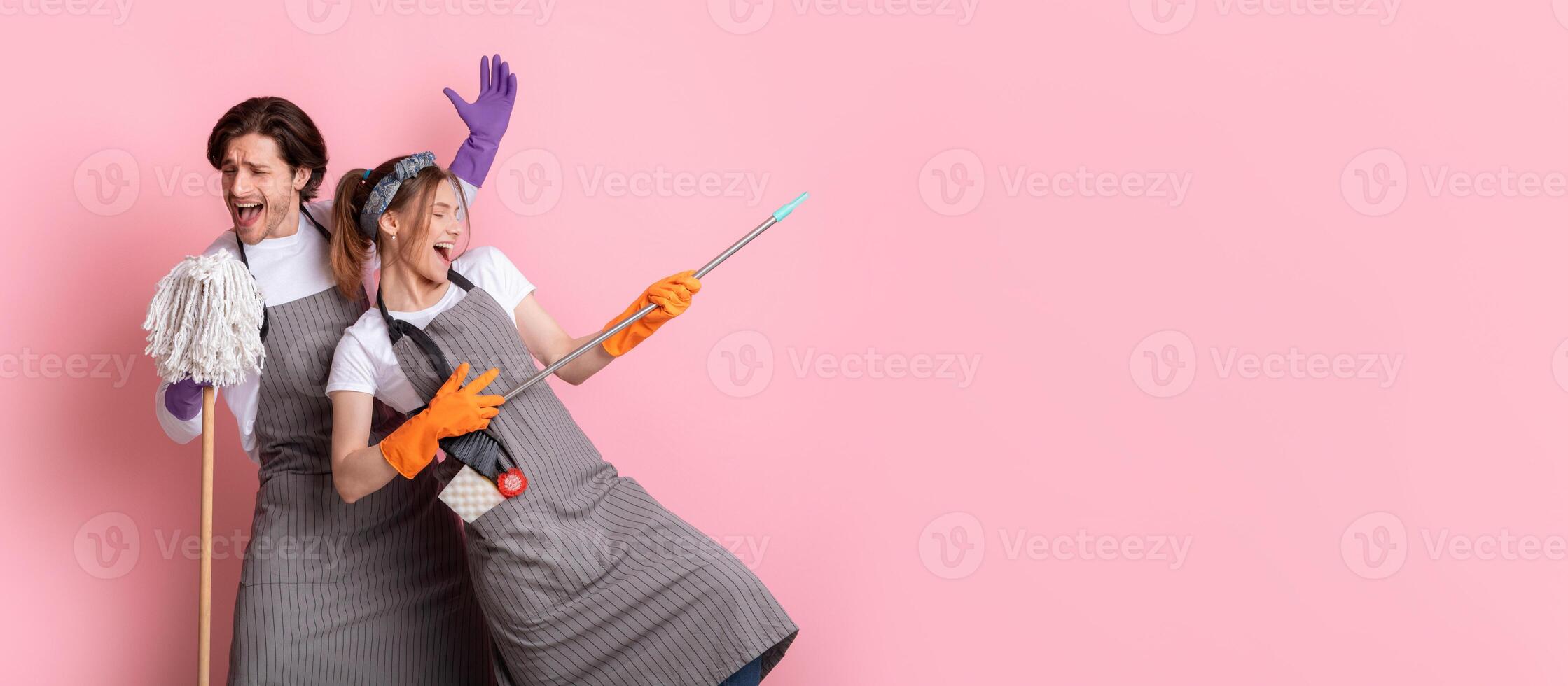 Young couple of janitors having fun during cleaning, fooling with mop and broom over pink background, panorama with copy space photo