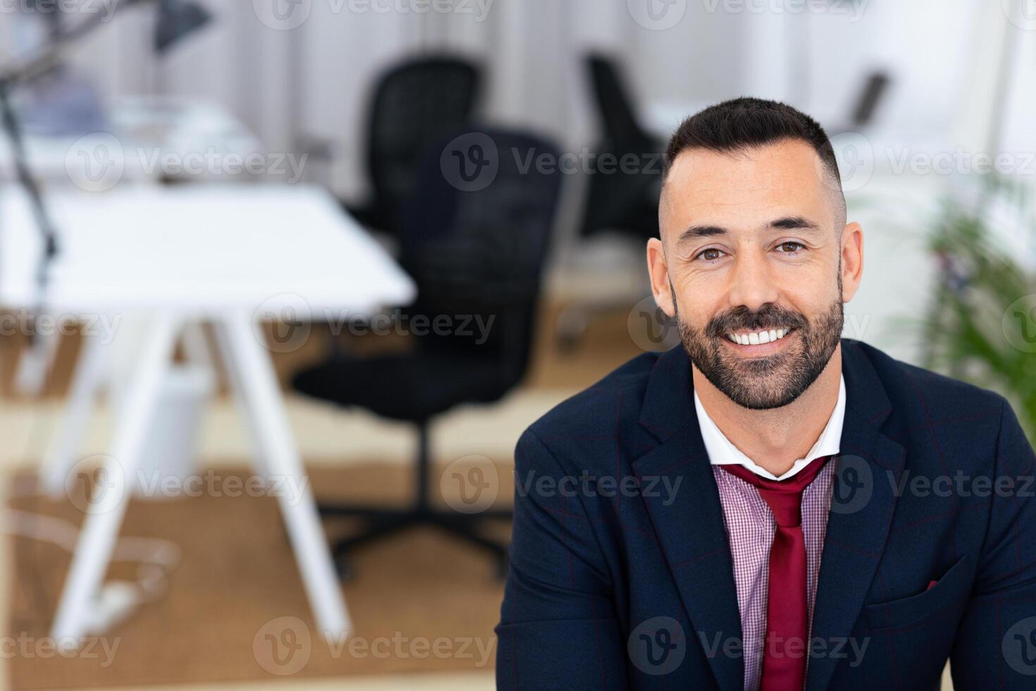 A well-dressed man with a neat beard and short hair smiles warmly while seated in a contemporary office. Behind him, stylish office furniture and a bright atmosphere enhance the environment. photo