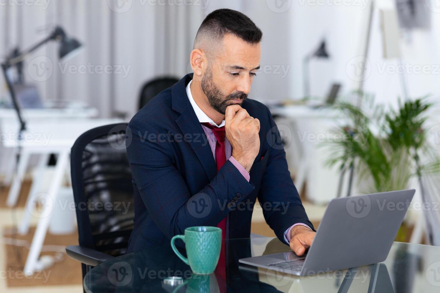 A man in a suit is focused on his laptop in a contemporary office. He appears to be deep in thought, holding his chin while typing, surrounded by various office elements. photo