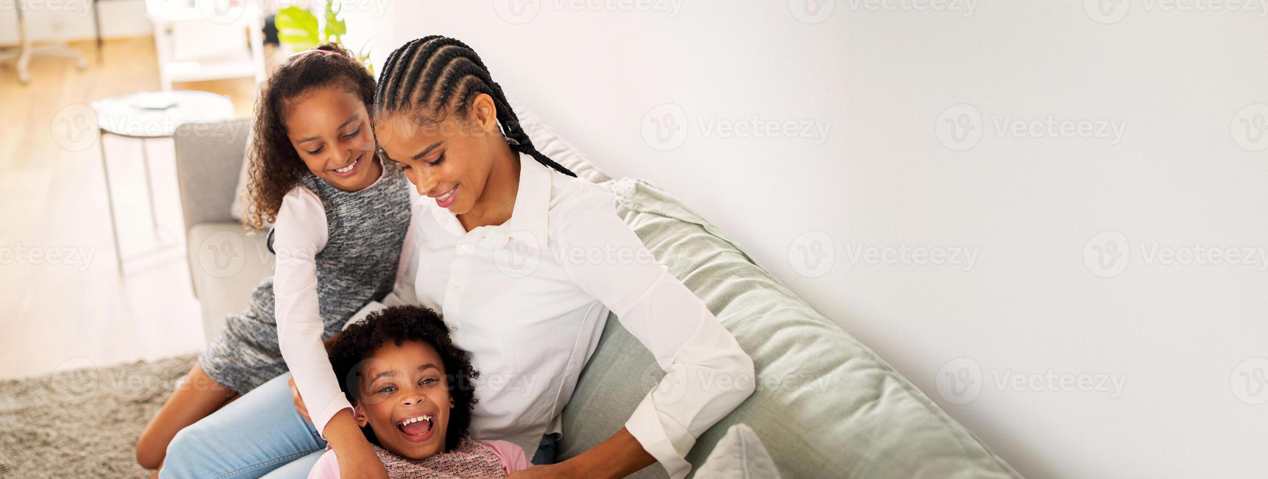 A black mother sits on her couch with her two daughters, laughing and playing together. The warm atmosphere in the living room highlights their joyful bonding time. photo