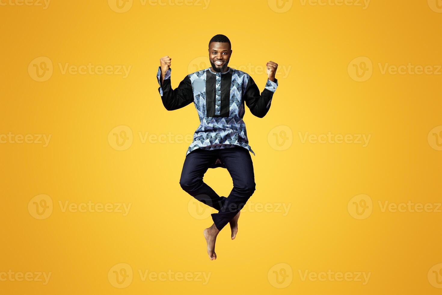 Emotional black guy showing happiness over pink studio background. Smiling african american man in colorful traditional clothes jumping up and posing, raising fists up, expressing emotions photo