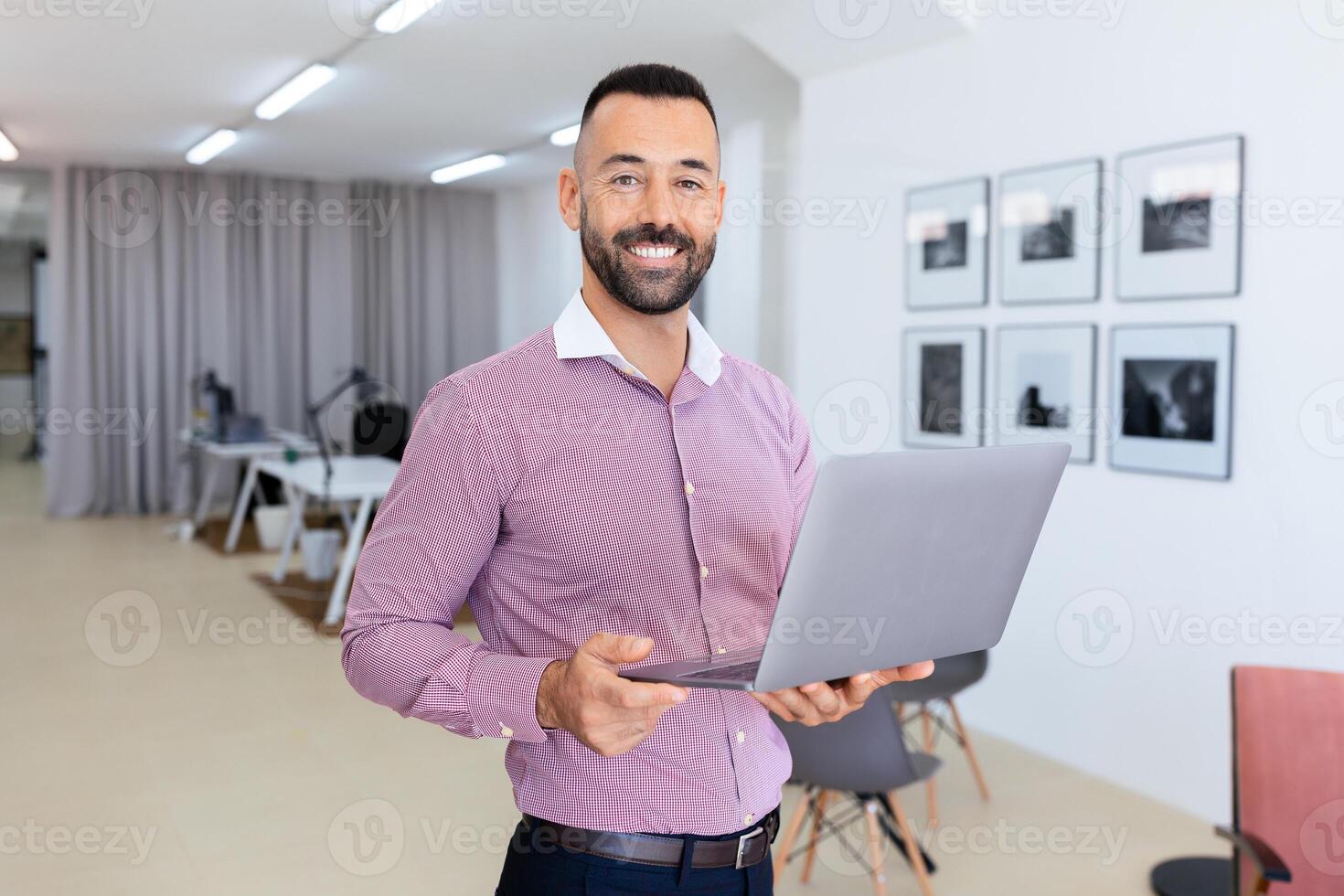 A well-dressed man stands in a contemporary office holding a laptop and smiling. The workspace features stylish furnishings and framed images on the wall. photo