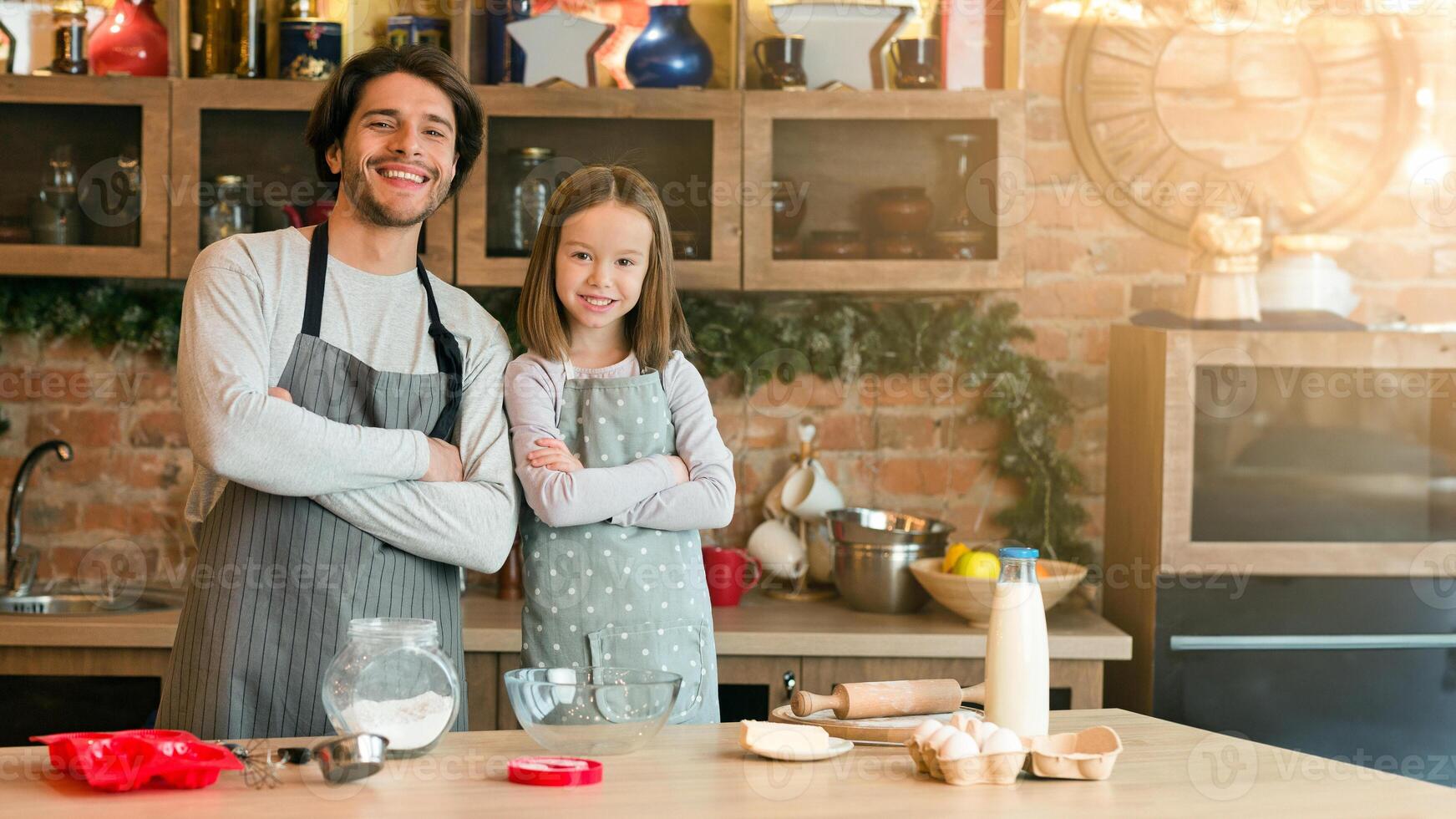 Young Handsome Dad And His Little Daughter In Aprons Posing In Kitchen While Preparing Pastry Together, Enjoying Homemade Food, Cooking Pie, Baking Sweets, Standing With Folded Arms, Smiling At Camera photo