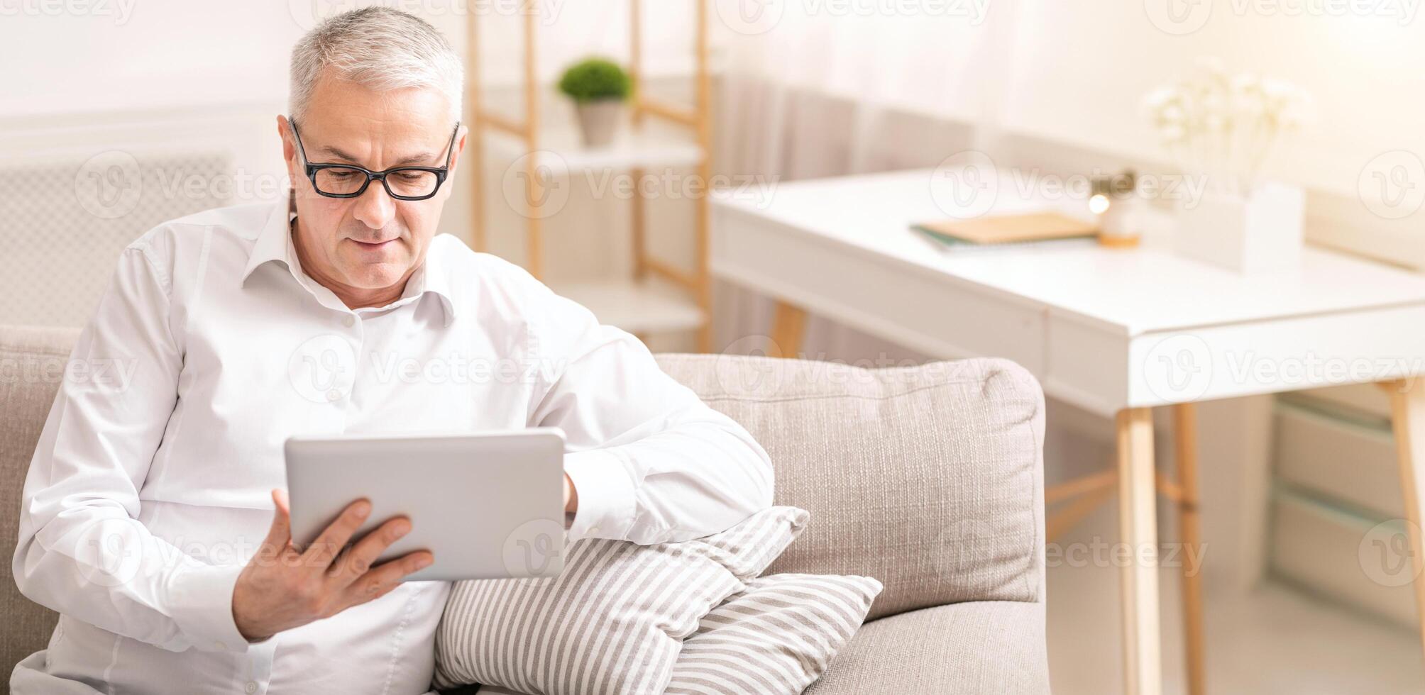 Reading News. Senior man in spectacles using digital tablet sitting on the couch at home, copyspace photo