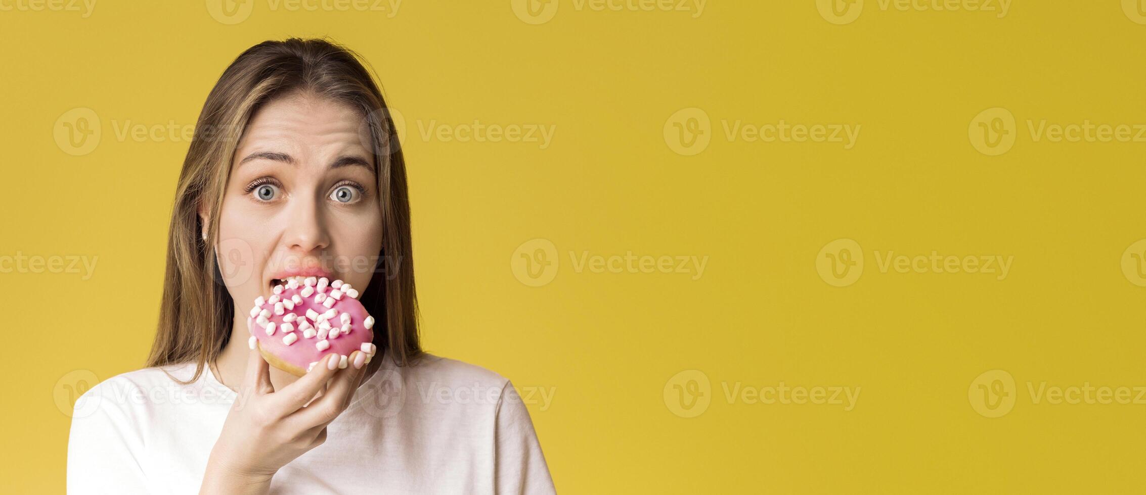 Young Lady Eating Donut, Preferring Unhealthy Food, Posing Over Yellow Background With Guilty Face Expression, Empty Space photo
