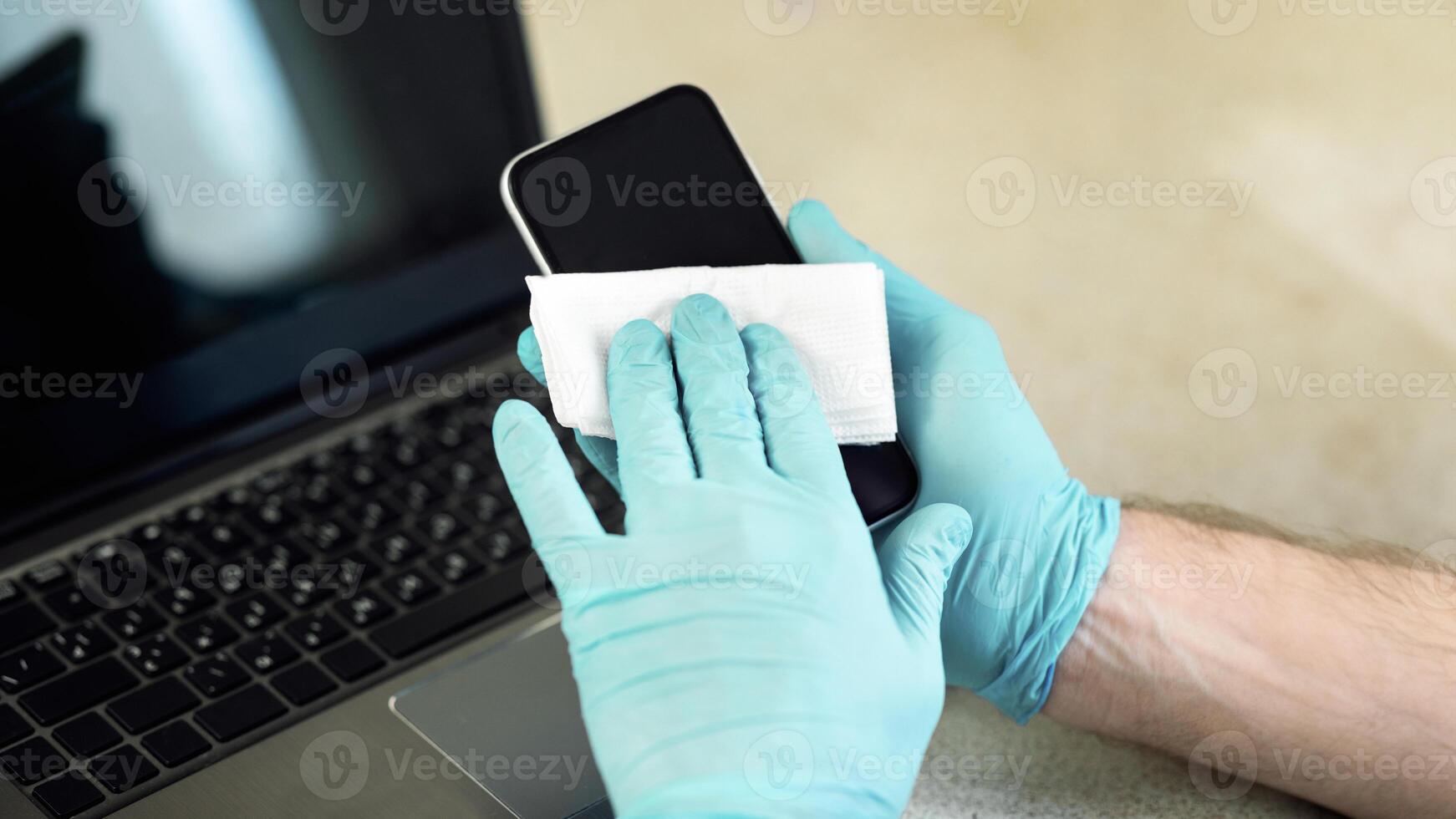 Unrecognizable Man Disinfecting Smartphone Before Use Sitting At Workplace, Wearing Protective Gloves Indoor. Coronavirus Protection And Hygiene. Cropped, Selective Focus photo