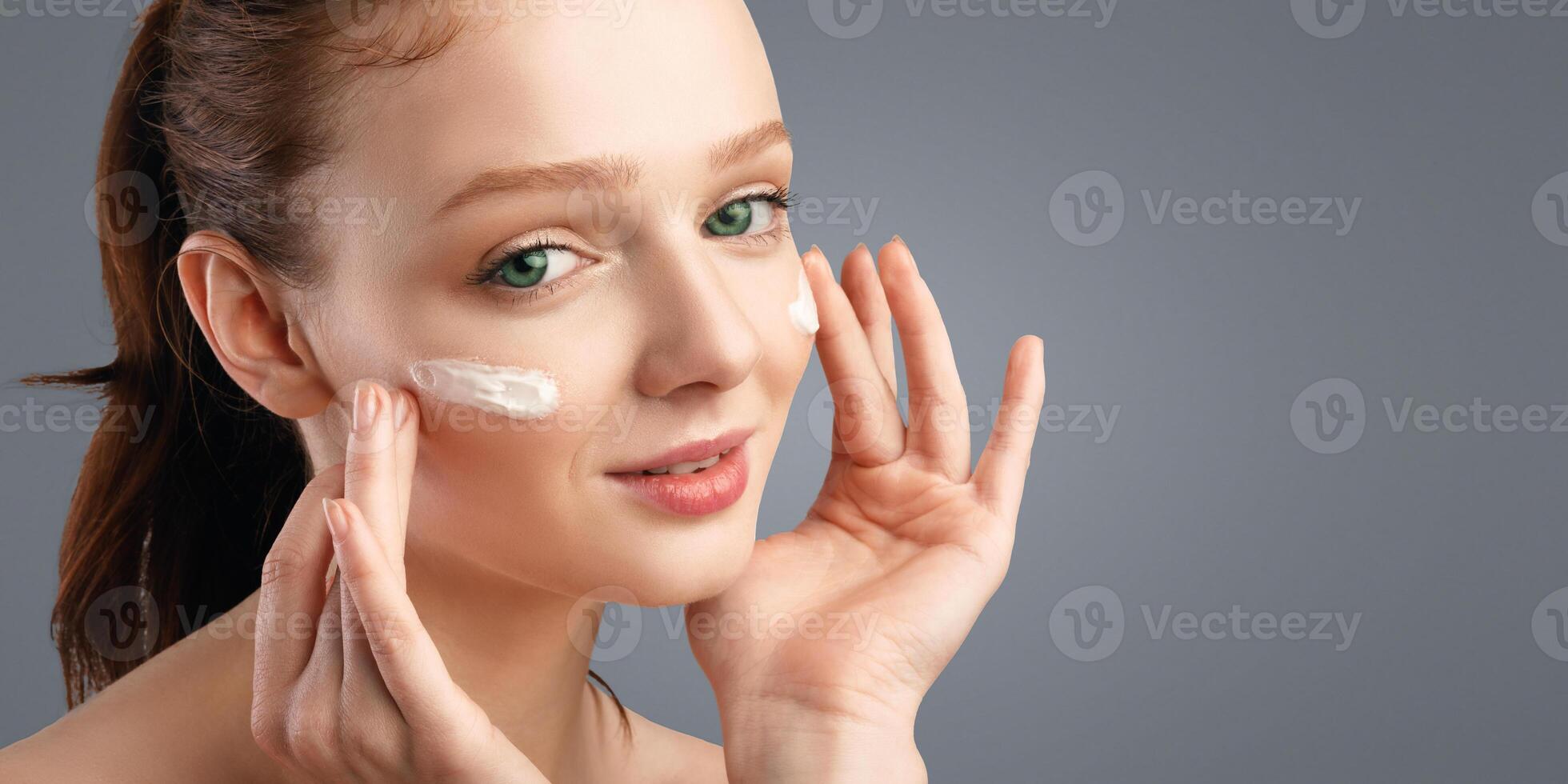Closeup Of Red-Haired Young Woman Applying Moisturizing Cream On Face Caring For Skin Smiling To Camera Standing Posing On White Background. Studio Shot. Skincare For Young Lady photo