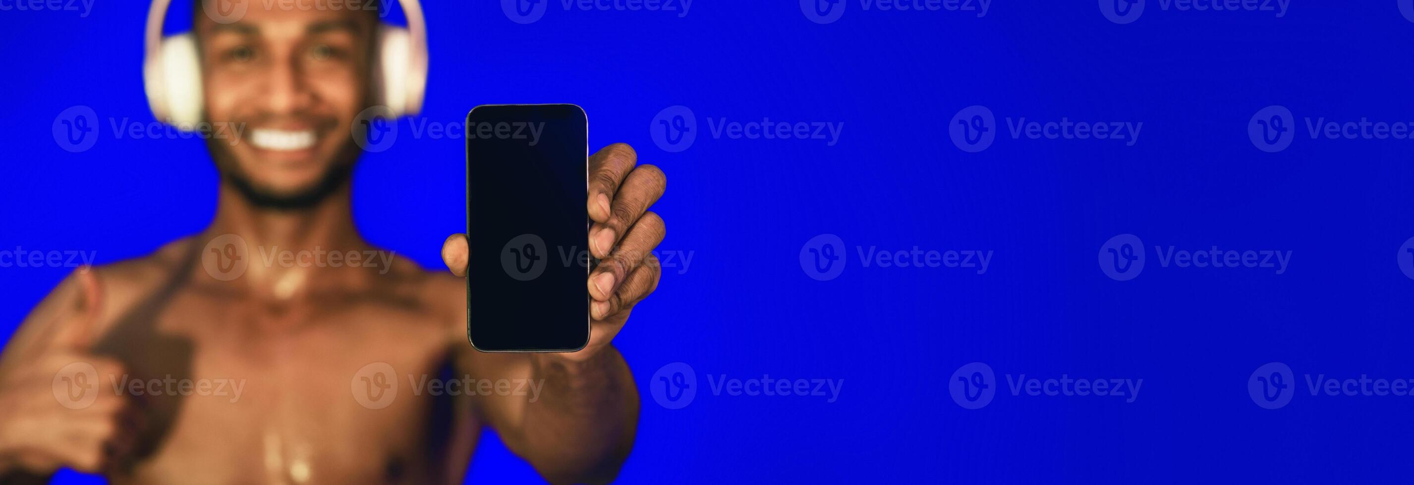 Sportsman With Headphones Showing Empty Phone Screen Gesturing Thumbs Up In Studio Over Blue Background. Mockup, Shallow Depth, Selective Focus photo