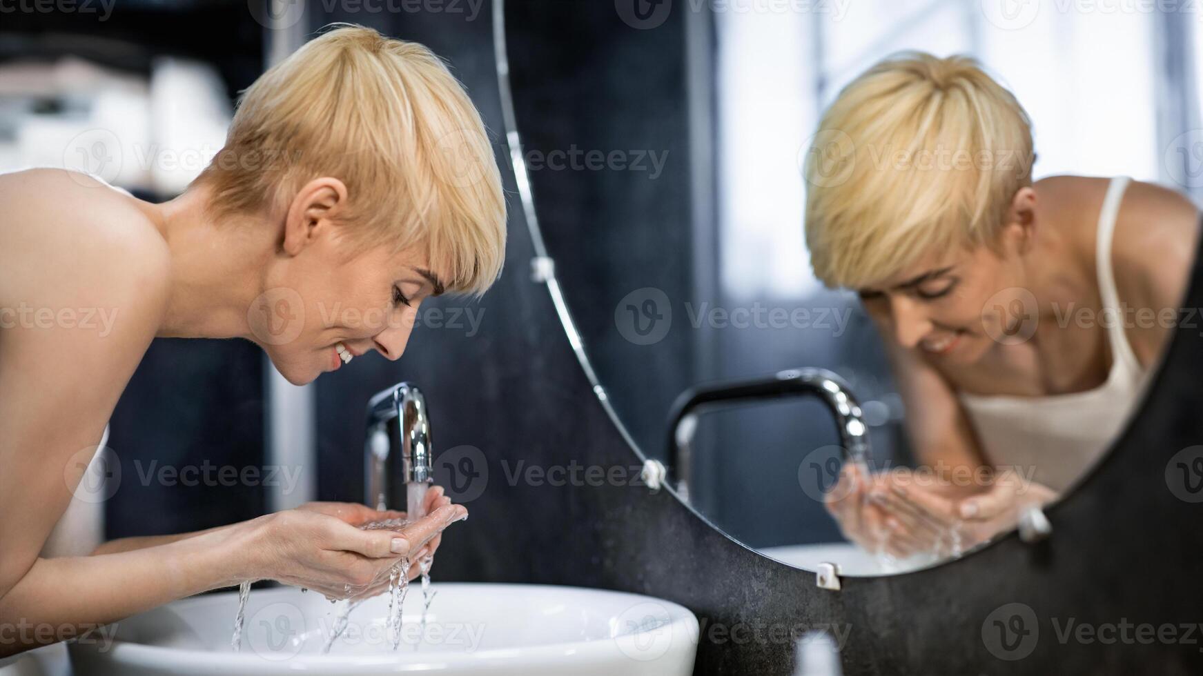 Cheerful Middle-Aged Lady Washing Face Standing In Bathroom. Hygiene And Skincare Routine. Selective Focus photo