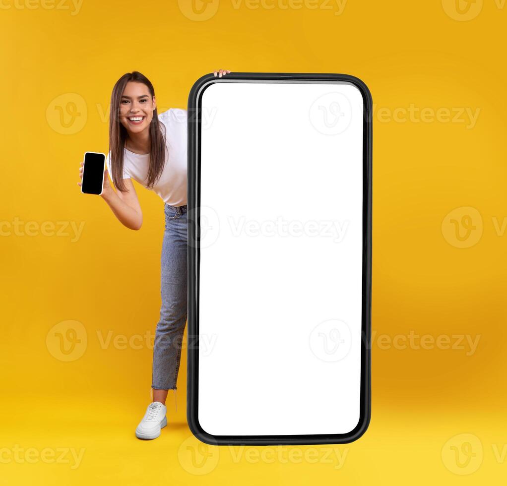 Full Body Length Of Cheerful Woman Peeking Out Standing Behind Big Smartphone With White Blank Screen, Excited Lady Holding Cell Phone Presenting New App Showing Copy Space For Website Design Mock Up photo