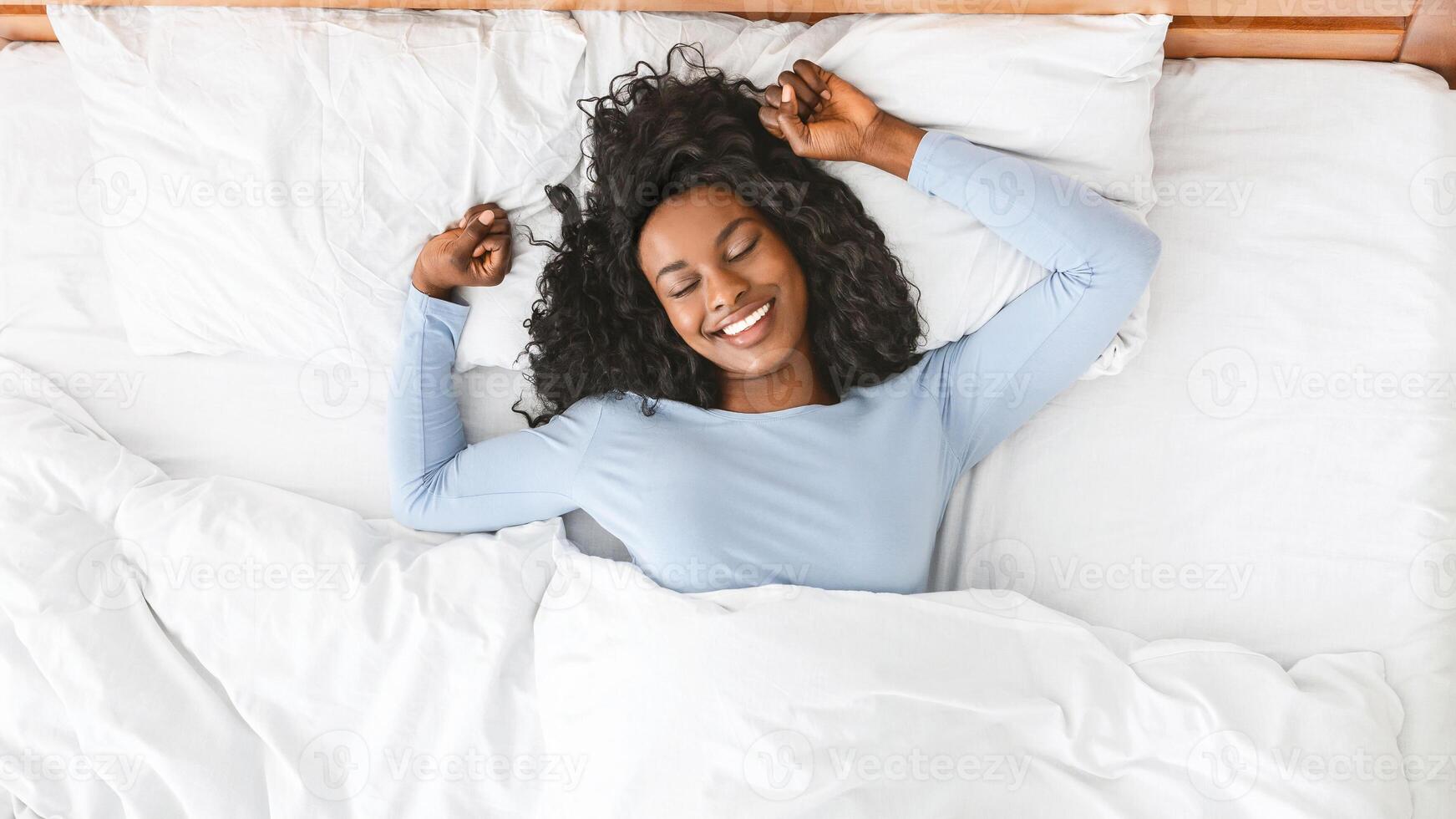 Young well-slept african woman stretching in bed, smiling with closed eyes, empty space, top view photo