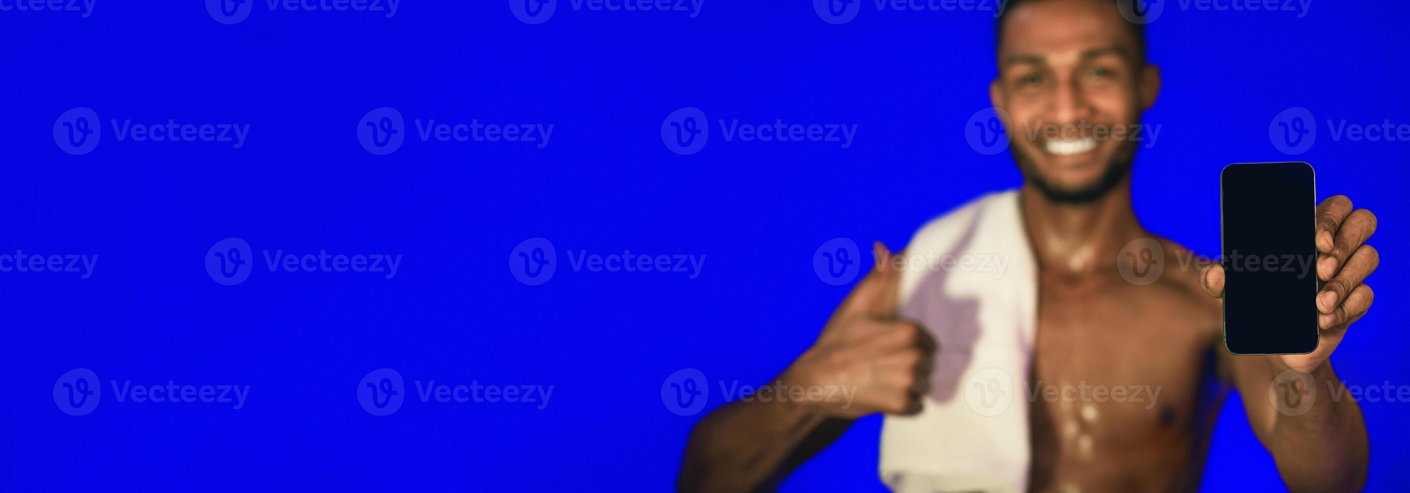 Workout Application. Athlete Guy Showing Smartphone Blank Screen Gesturing Thumbs-Up Standing On Blue Studio Background. Mockup, Shallow Depth photo