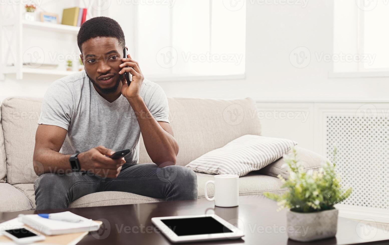 Young happy african-american man watching tv and talking on smartphone pointing with remote controller on tv-set, sitting on the couch, copy space photo