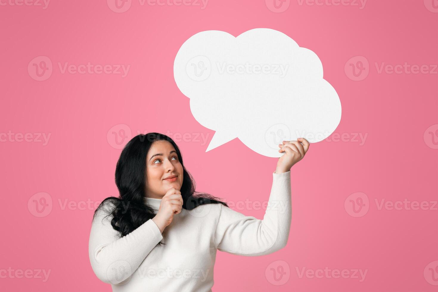 Pensive plus size woman holding and looking at blank speech bubble over her head, standing on pink background photo