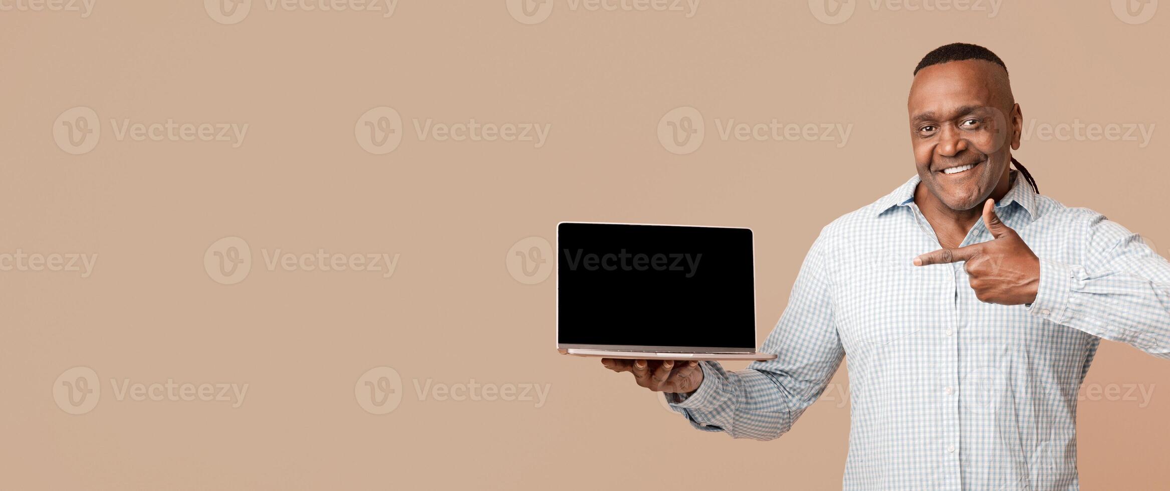 Awesome website. Middle aged positive afro man pointing at blank screen of laptop, orange studio background photo