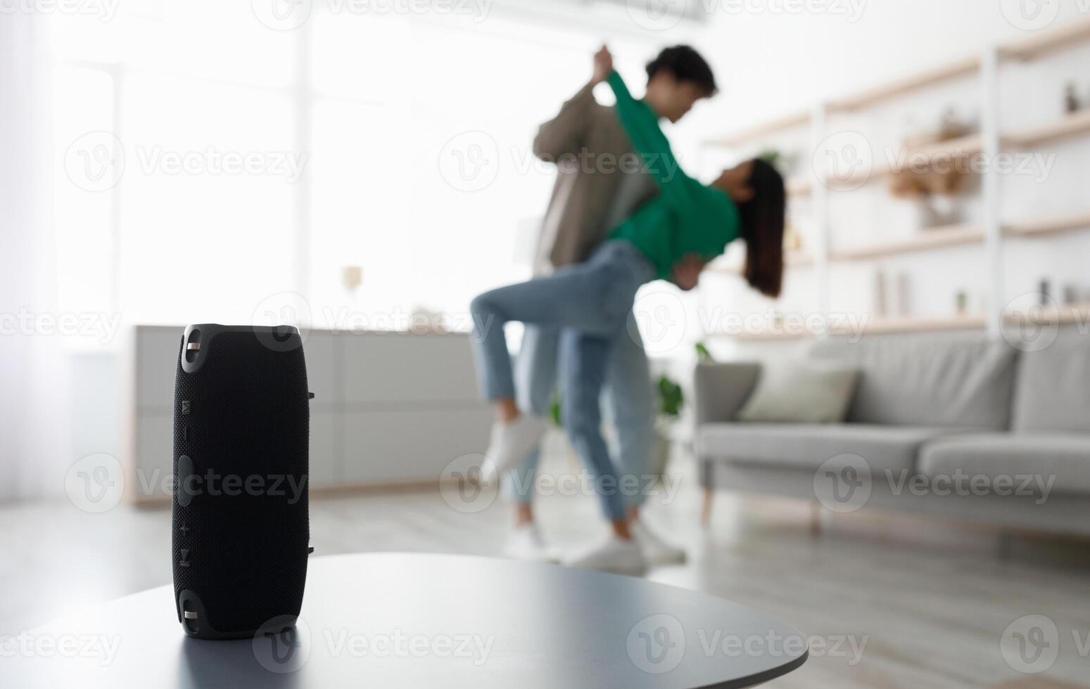 Millennial Asian couple dancing to popular music at home, selective focus on portable wireless speaker on table, copy space. Young family moving to their favorite song, using modern device photo