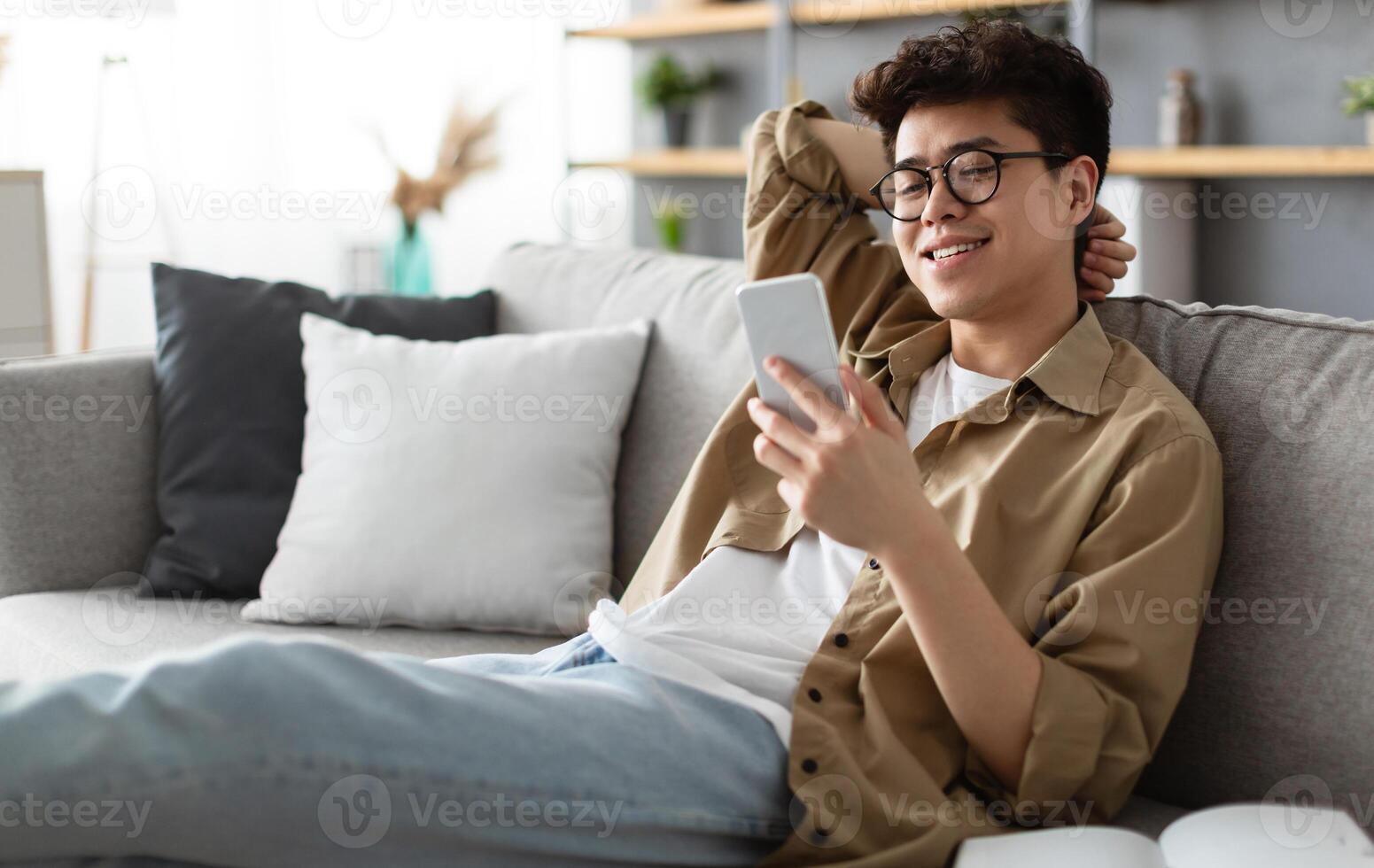 Enjoying Weekend. Handsome Asian Man In Glasses Using Smartphone While Sitting Leaning On Couch At Home, Happy Guy Relaxing In Living Room, Browsing Internet Or Messaging With Friends, Selective Focus photo
