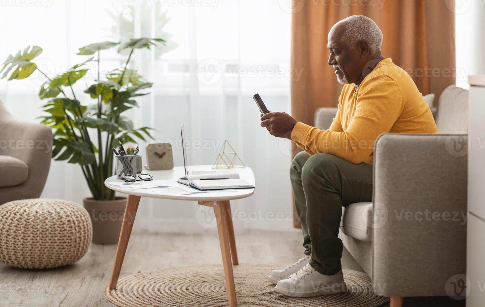 Side View Of Mature Black Man Using Smartphone Texting Sitting At Laptop Working At Home. Male Freelancer Browsing Internet Via Cellphone Or Using Mobile Application. Technology And Gadgets photo