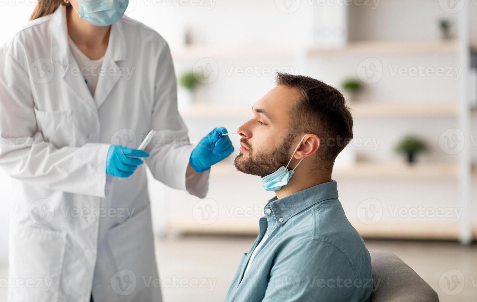 Covid diagnostic procedure. Doctor making nasal PCR test for young Caucasian man, using sterile swab stick at home. Millennial guy undergoing coronavirus analysis, checking health photo