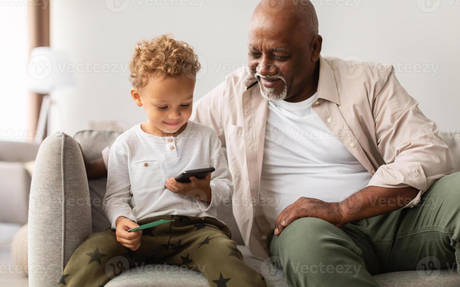 negro abuelo y pequeño nieto utilizando Teléfono móvil jugando móvil juego juntos sentado a hogar. niño chico hojeada Internet y utilizando nuevo solicitud en teléfono inteligente gasto hora con abuelo foto