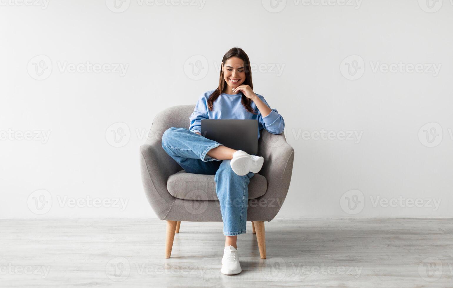 Millennial Caucasian lady in casual wear using laptop, sitting in armchair, having online conference against white studio wall. Young woman communicating online, taking part in webinar photo