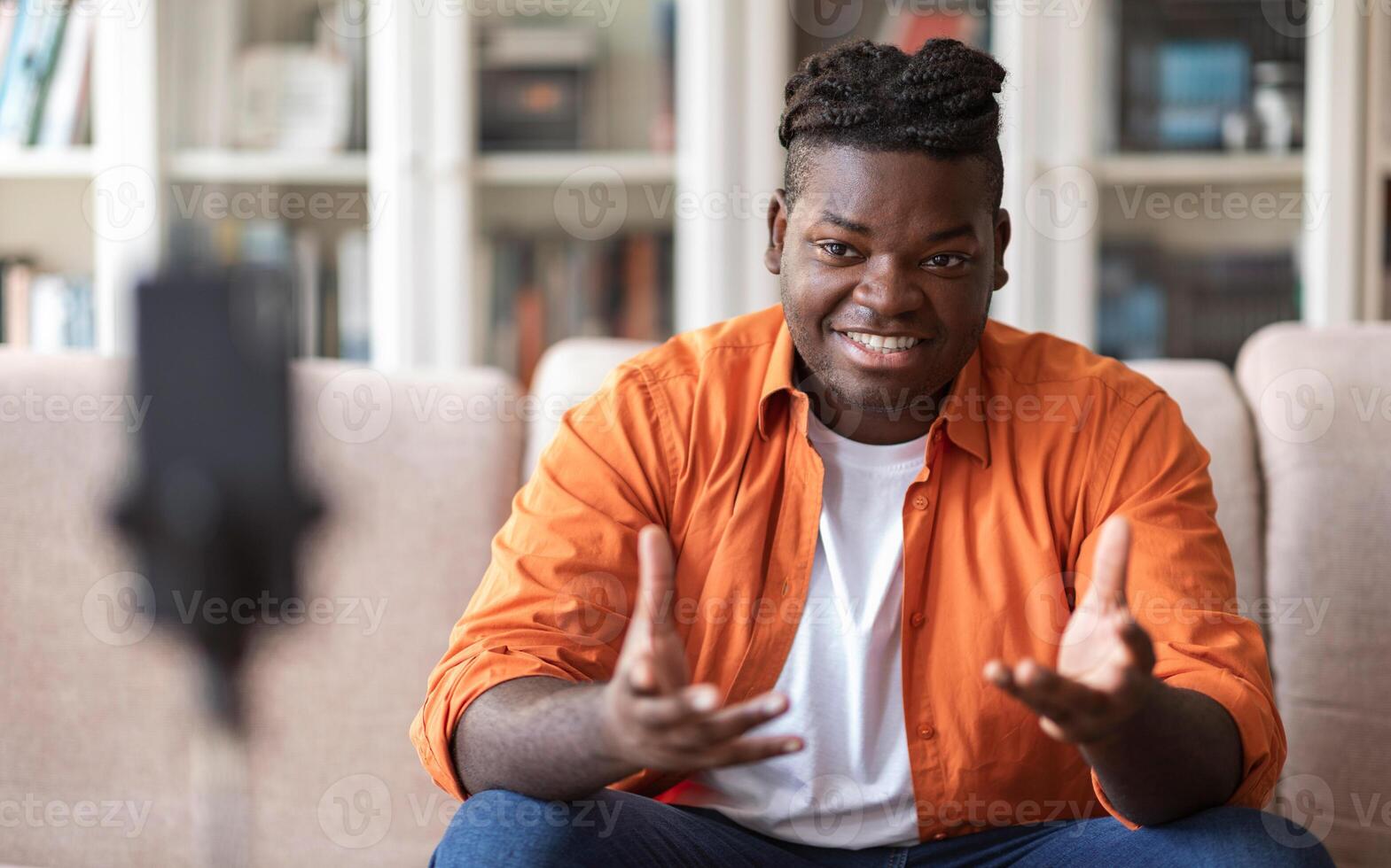 Blogging concept. Young African American chuby guy blogger in casual clothes streaming online or recording tutorial for social media network, looking at the mobile phone on a tripod and smiling photo