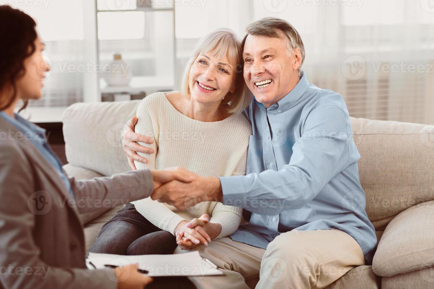 Sealing A Deal. Happy elderly couple sitting on couch and shaking hands with young female financial advisor photo