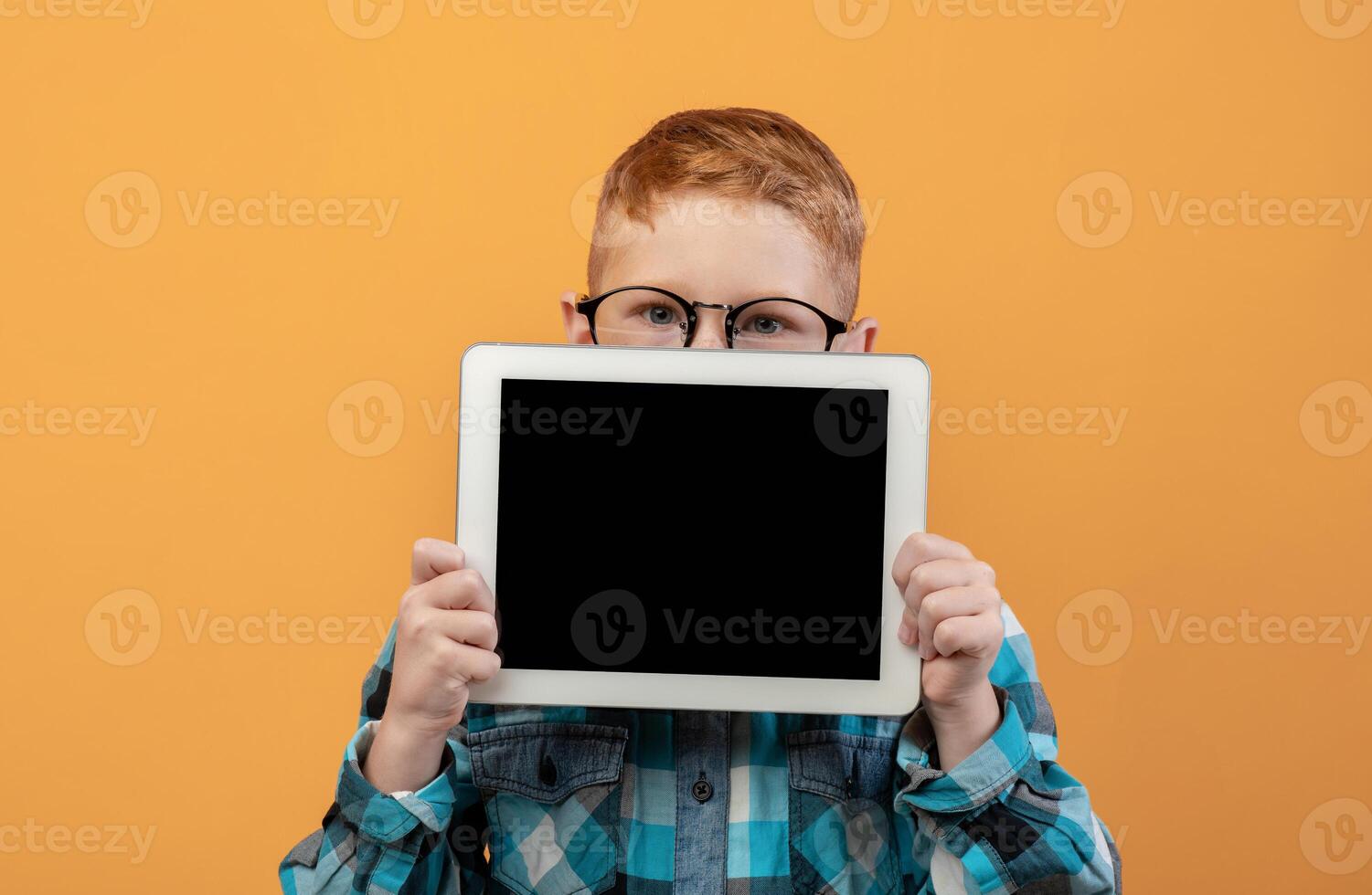 Ginger boy in eyeglasses hiding behind digital tablet with empty screen over yellow studio background. Cute redhead schoolboy showing pad, mockup, advertisement for cute application photo