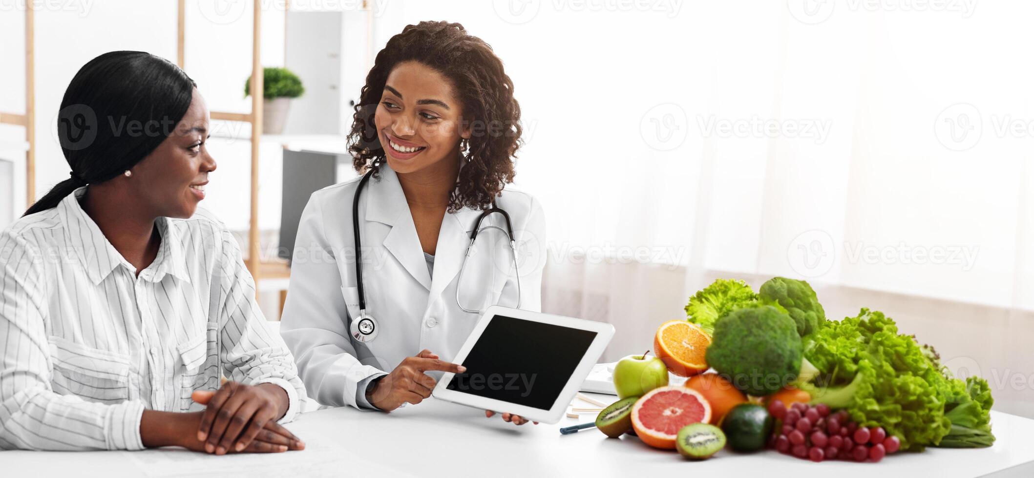 Friendly female dietologist showing black lady patient empty digital tablet screen in her office, technologies in medical care photo