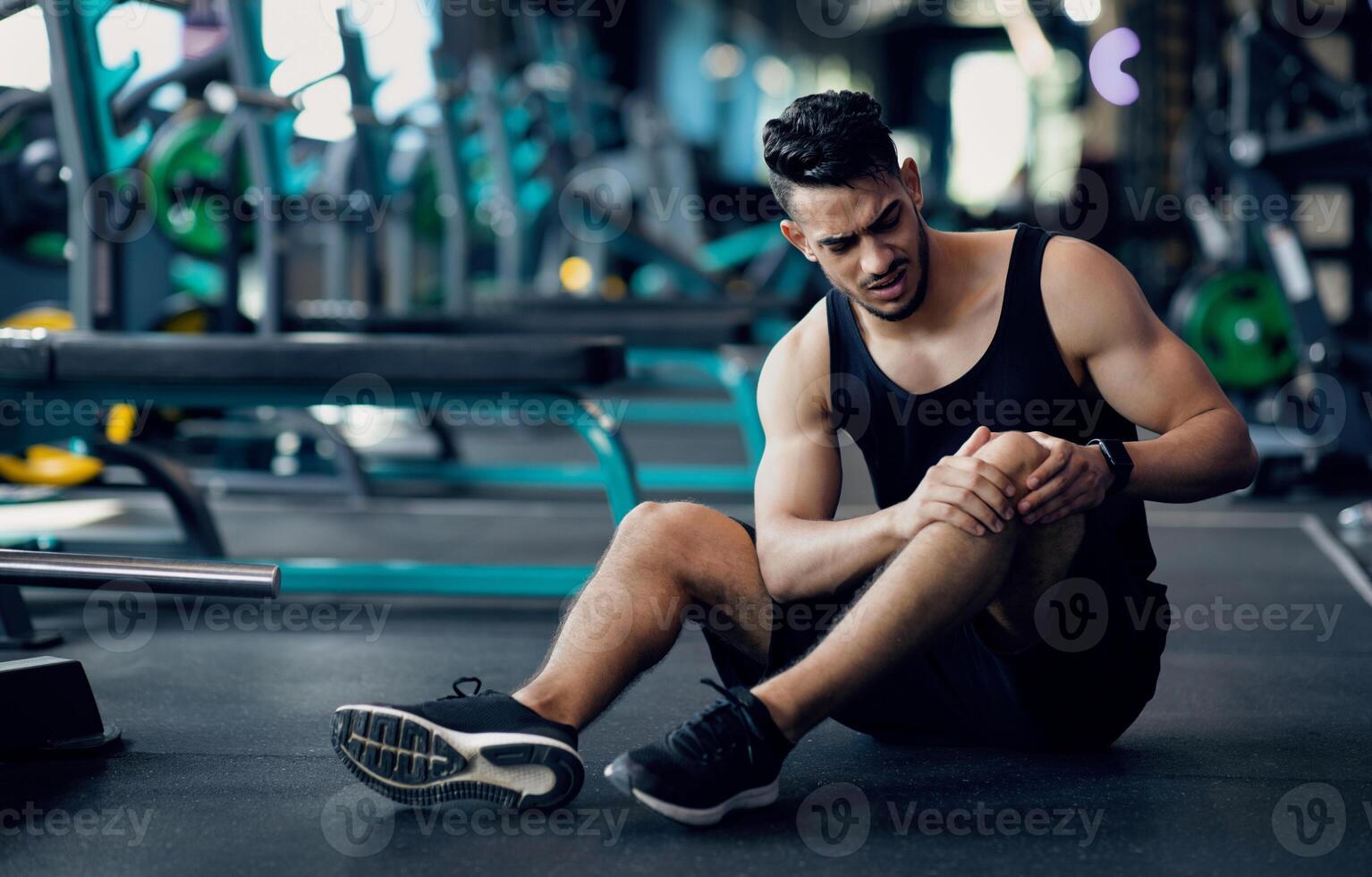 Sport Trauma. Young Arab Male Athlete Touching His Hurted Knee At Gym, Upset Middle Eastern Guy Sitting On Floor And Massaging Sore Injured Leg After Workout Training In Fitness Club, Free Space photo
