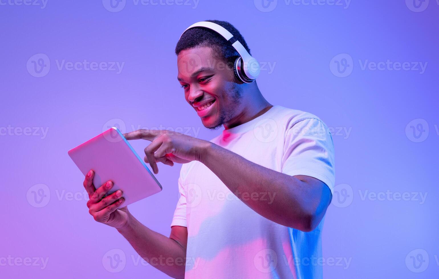 Interesting App. Portrait Of Excited African Guy In Wireless Headphones Using Digital Tablet, Millennial Black Man Playing Games Or Browsing Internet While Standing Under Purple Neon Light photo