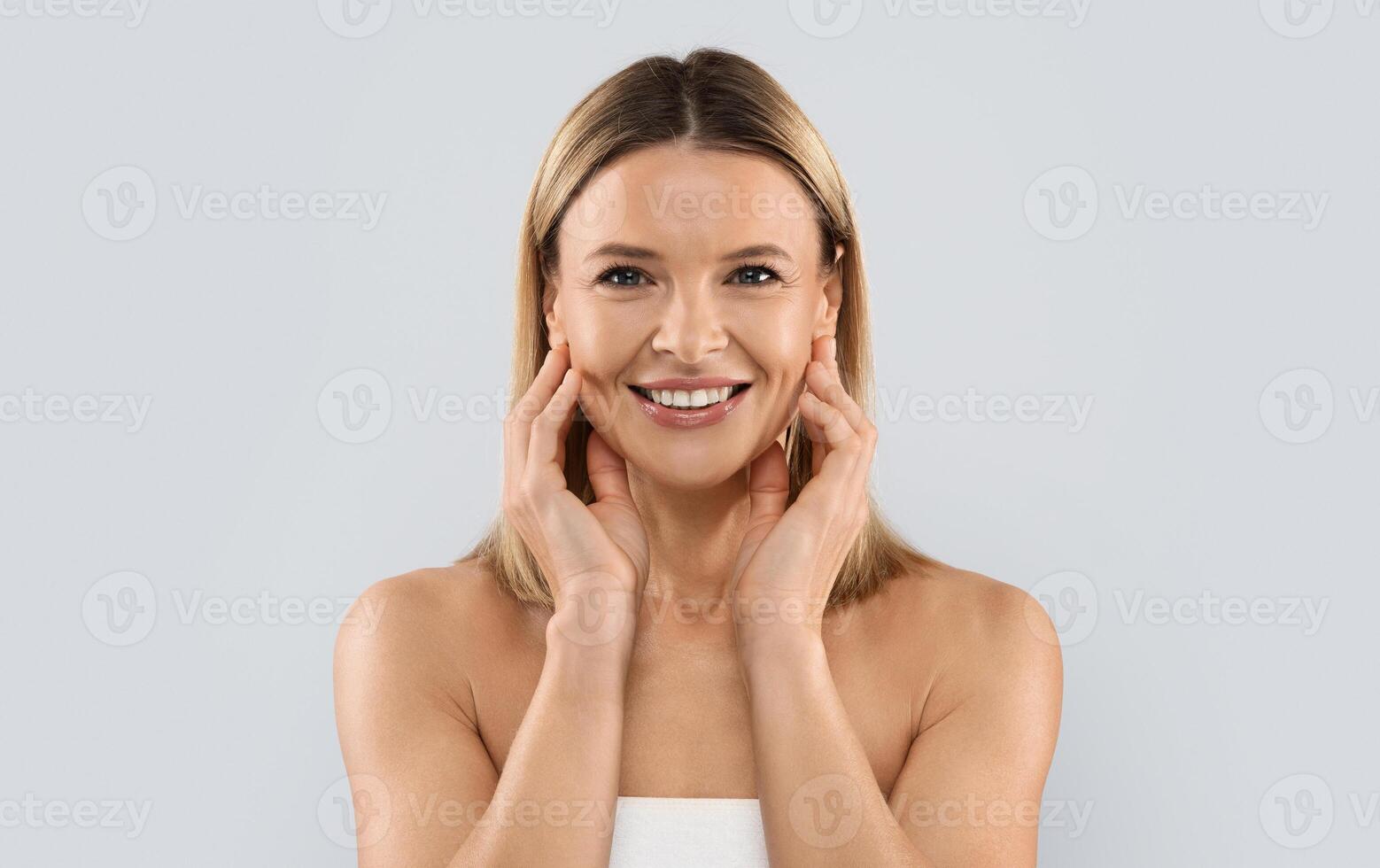 Face injections concept. Pretty blonde middle aged woman wrapped in white bath towel touching her face skin and smiling, enjoying results of face care treatment, grey studio background photo