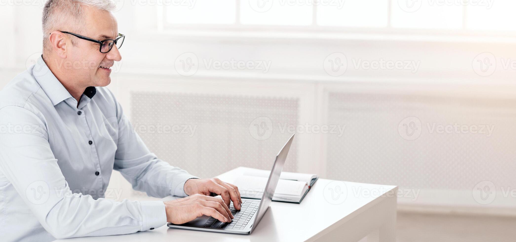 Home Office Concept. Smiling Mature Man In Eyeglasses Typing Report On Computer Laptop Sitting At Table, Copyspace photo
