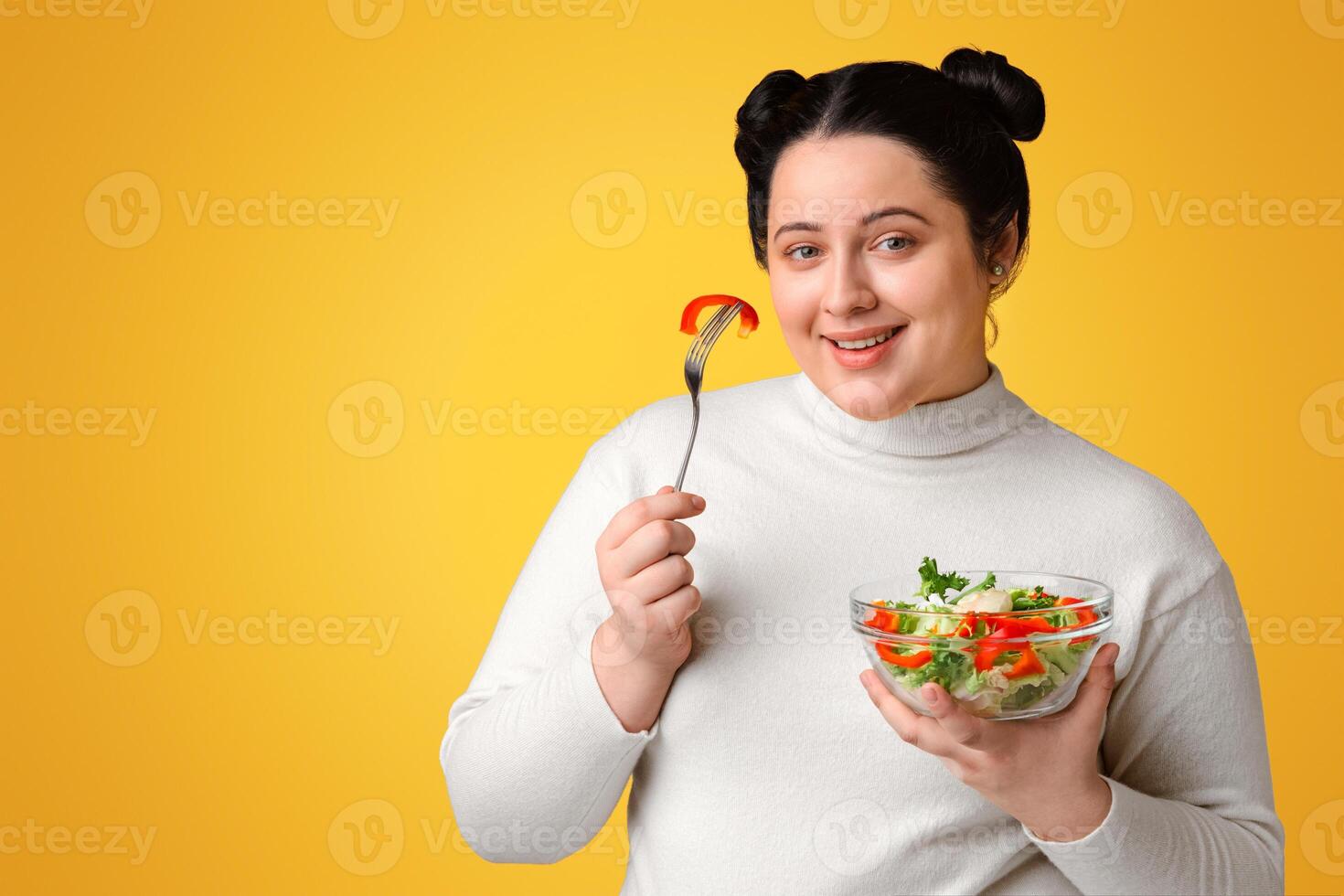 Healthy Nutrition. Happy Young Plumb Woman Eating Fresh Vegetables Salad, Posing Over Yellow Background In Studio With Bowl And Fork, Empty Space photo