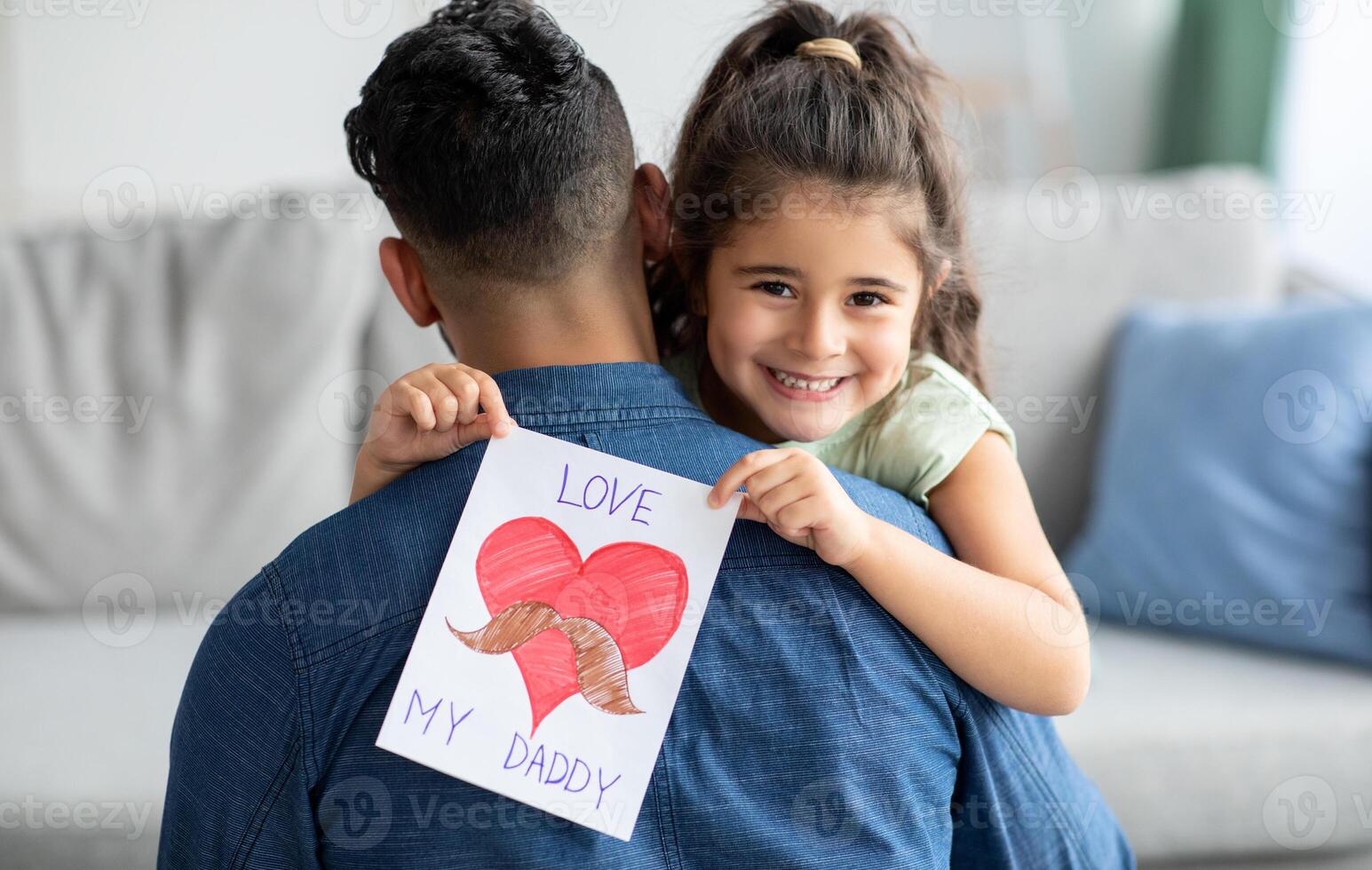 Father's Day Concept. Smiling little girl embracing dad and holding greeting card with drawn big red heart, greeting daddy with holiday, cheerful female child looking at camera, closeup shot photo