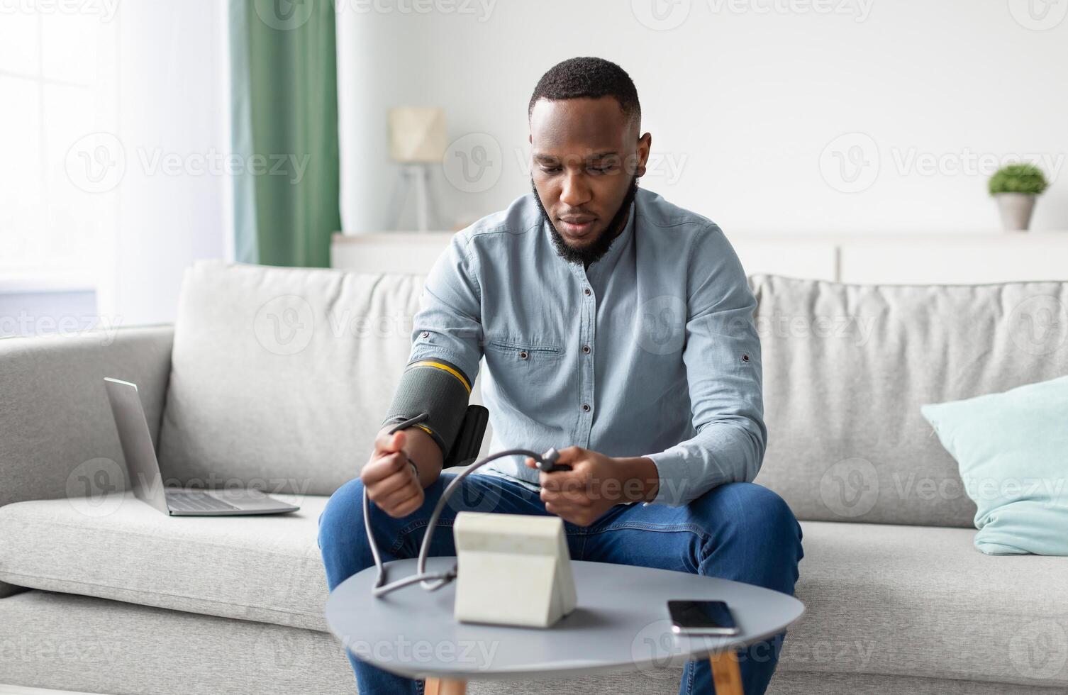 Hypertension. African Man Measuring Arterial Blood Pressure Having Health Problem Sitting On Sofa At Home. Hypotension And High Pressure Issue Concept. Selective Focus photo